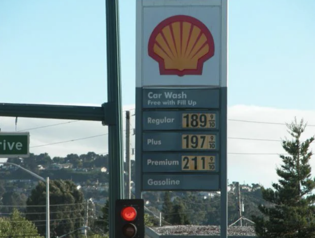 A Shell gas station sign shows prices for different fuel types. Regular is priced at $1.89 9/10, Plus at $1.97 9/10, and Premium at $2.11 9/10 per gallon. A green street sign and a traffic light on red are in the foreground, with trees and hills in the background.