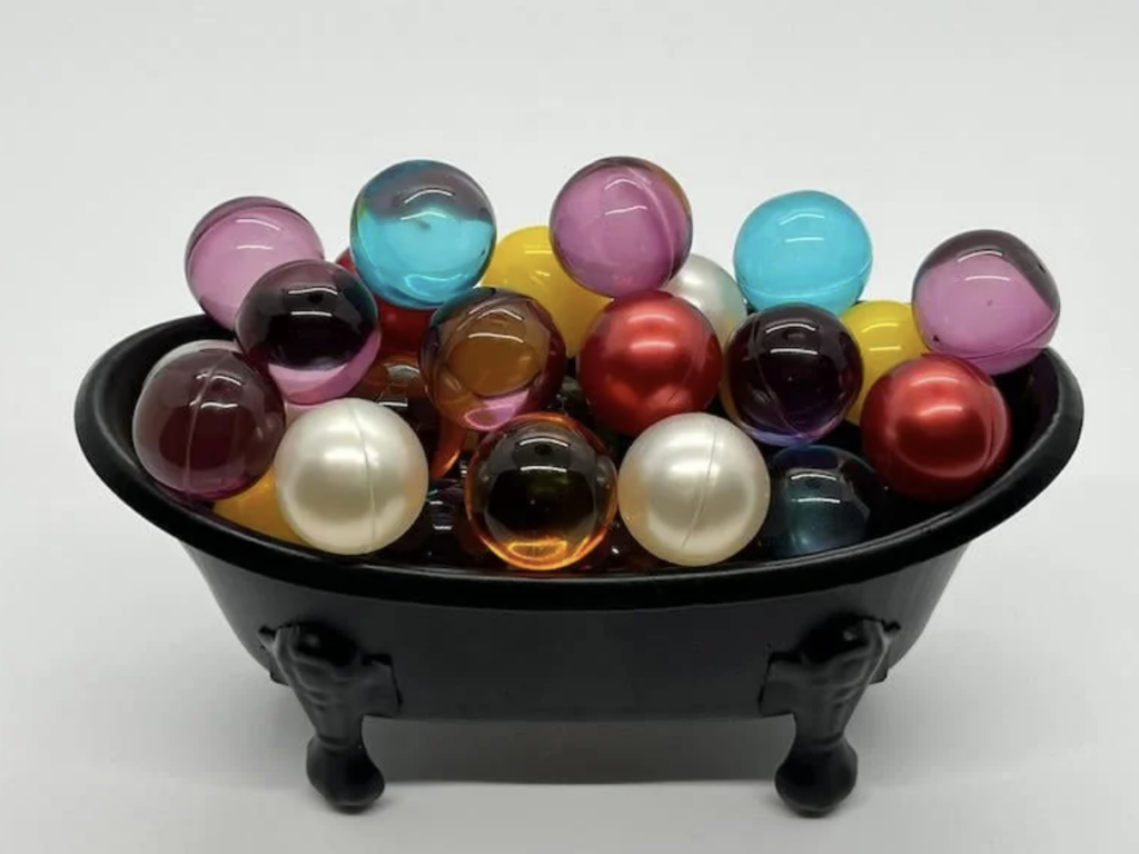 A small black bathtub is filled with a variety of colorful, translucent marbles and shiny white pearls. The marbles come in shades of pink, blue, yellow, red, and clear. The bathtub stands on four legs and is set against a plain white background.