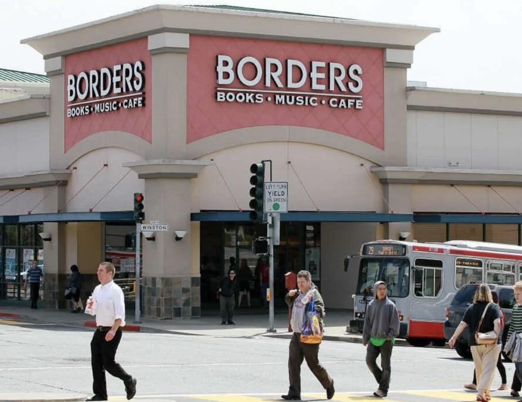 A large Borders bookstore with the signage "BOOKS, MUSIC, CAFE" on the building's facade. People are walking in front of the store, and a bus is stopped near the corner. The scene is set in an urban area with traffic lights and crosswalks in view.