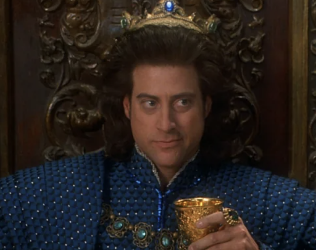 A man with long, dark brown hair and a crown sits on an ornate, carved wooden throne. He is wearing a blue outfit adorned with intricate designs and jewels. He holds a golden chalice in his left hand and appears to be looking to his right with a slight smile.