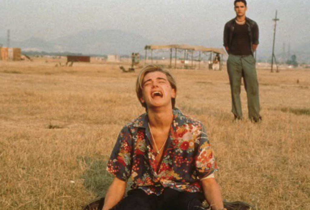 A young man in a brightly colored floral shirt sits on dry grass, crying out in despair with his mouth open and head tilted back. Another man stands in the background, wearing a dark shirt and green pants, looking on with a serious expression. The scene is set outdoors.