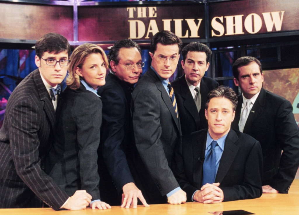 A group of seven people in formal attire stand and sit around a desk with "The Daily Show" sign in the background. Some individuals are making serious expressions while others are adopting humorous poses.