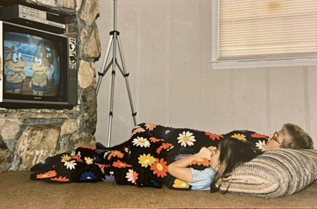 Two children lie on the floor under a black blanket with colorful flower patterns, watching a cartoon on an old tube television set. The TV is placed in a stone fireplace. The room has a beige carpet and walls with a window covered by blinds in the background.