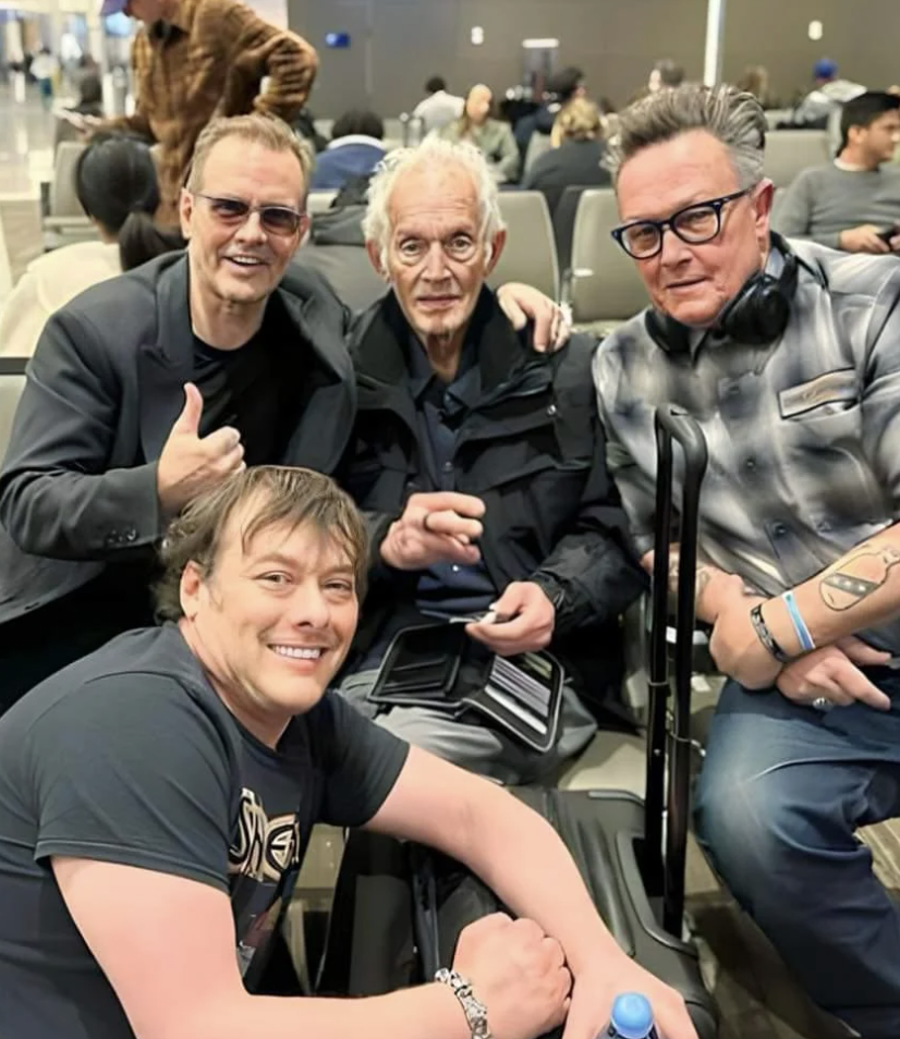 Four men are posing together and smiling indoors. One man, standing behind the rest, is giving a thumbs-up gesture. Two of the men are seated, with one holding a bottle of water and wearing headphones around his neck. The fourth man crouches in front with a broad smile.