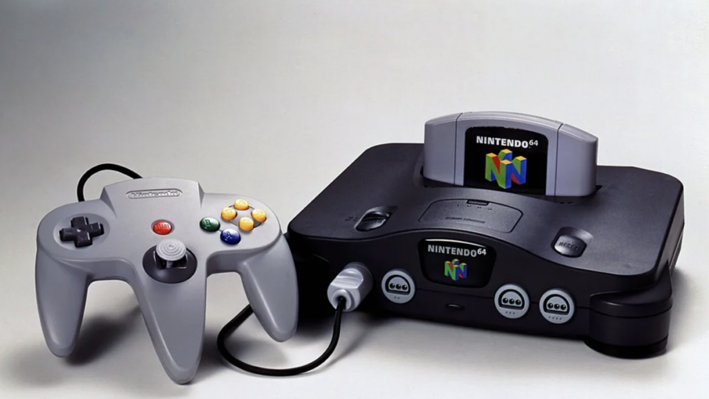 A black Nintendo 64 gaming console with a gray controller connected by a cord. The console has a game cartridge inserted, displaying the Nintendo 64 logo. The controller features an analog stick, a directional pad, and colorful buttons.