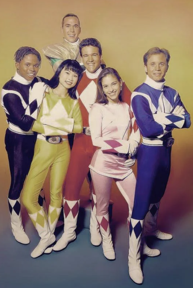 Six people dressed in Power Ranger costumes stand together against a gradient background. They wear helmets with matching suits in colors black, yellow, blue, red, pink, and green, and pose confidently with smiles and folded arms.