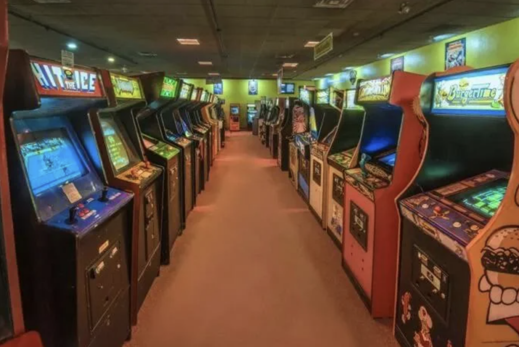 A dimly lit arcade filled with vintage game machines lined up in neat rows. Each machine has a brightly lit screen displaying various game titles and vibrant artwork on their cabinets. The floor is carpeted, and the ambiance is retro and nostalgic.