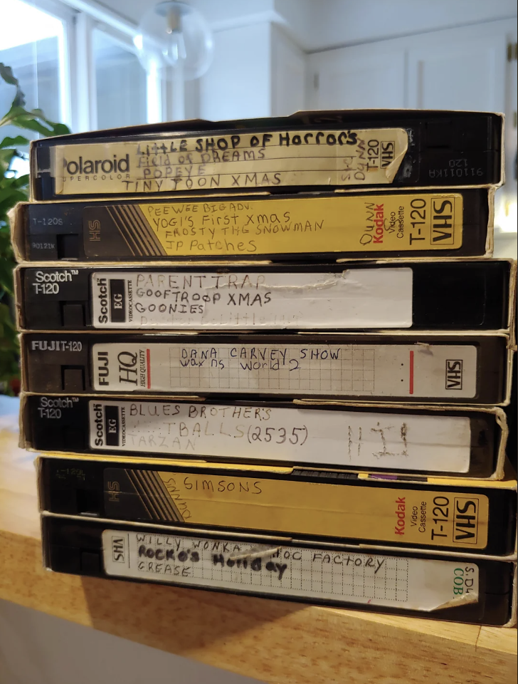 A stack of VHS tapes, each labeled with different titles. Labels include "Little Shop of Horrors," "Radio Dreams," "The Simpsons," "Blues Brothers," and others. The tapes are various brands, including Scotch and Polaroid, and are stacked on a wooden surface.