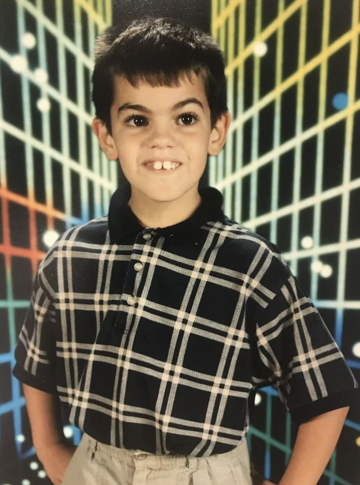 A young boy with short dark hair smiles while posing for a school photo. He is wearing a black and white plaid shirt and light-colored pants. The background features a colorful grid pattern with lines forming a perspective effect, accented by white dots.