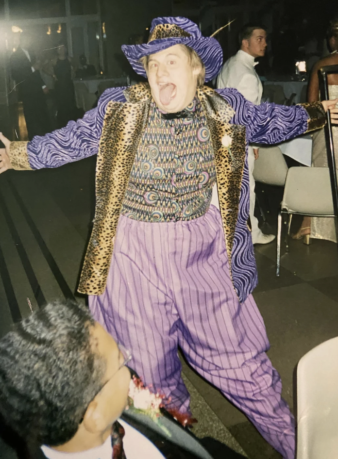 A person in a vibrant, eclectic outfit poses enthusiastically with arms outstretched. They wear a purple-patterned hat, a leopard-print jacket, a colorful patterned shirt, and baggy purple pants. The scene appears to be a festive event with other attendees visible in the background.