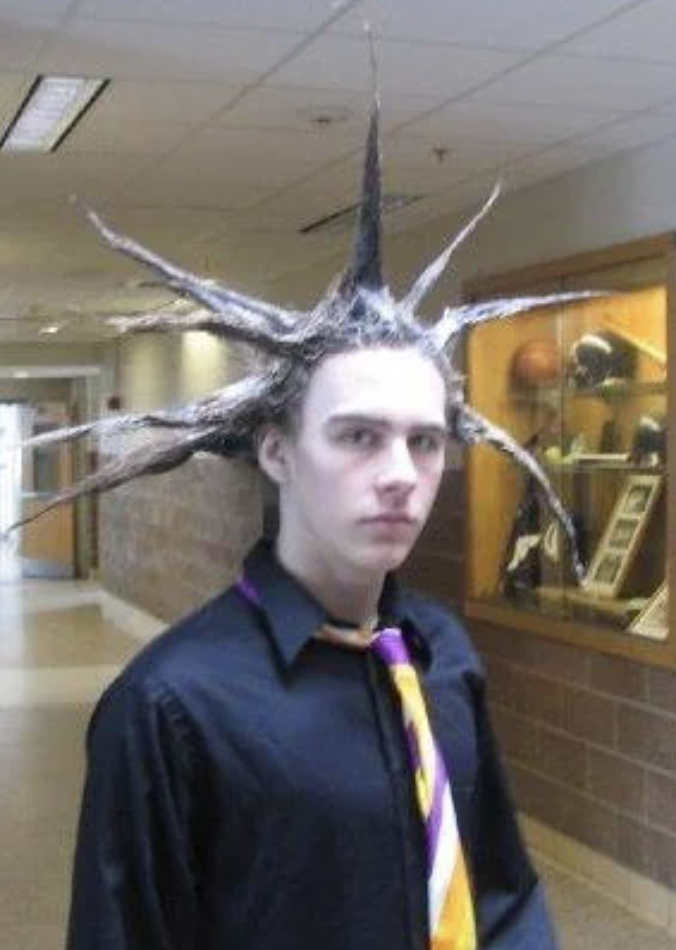 A person with light skin wearing a black shirt and a multicolored striped tie. Their hair is styled into multiple long, pointed spikes extending outward in all directions. They are standing in a hallway with display cases in the background.