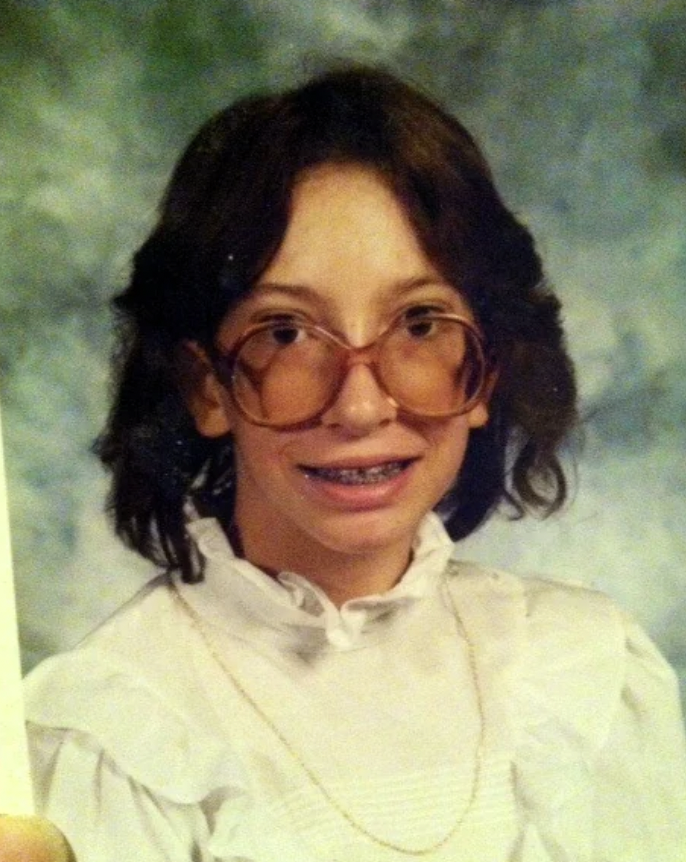 A young person with shoulder-length brown hair, wearing large brown glasses and braces, is smiling at the camera. They are dressed in a white ruffled shirt paired with a thin gold necklace. The background is a blurred mix of blue and green tones.