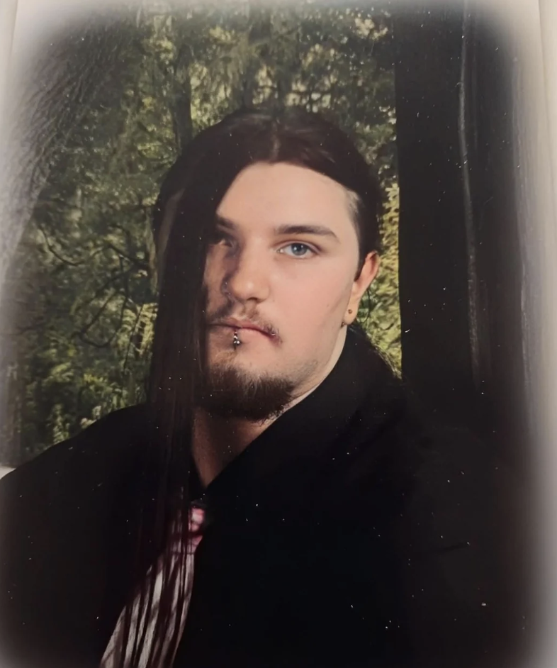 A person with long dark hair, a beard, and a lip piercing stares intently at the camera. They are dressed in dark clothing, and the background appears to be a blurred nature scene with verdant greenery. The image is softly focused at the edges.