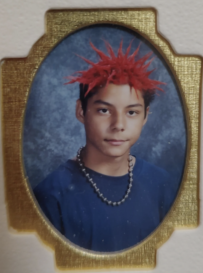 A framed portrait of a young person with bright red spiky hair, wearing a dark blue shirt and a large silver bead necklace. The background is a mottled blue and the frame is ornate and golden. The individual is looking directly at the camera with a neutral expression.