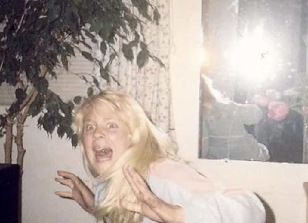 A person with long blonde hair and a surprised expression is standing indoors, hands raised near their face. They appear to be illuminated by a flash, with a potted plant and a window with curtains in the background. The photo's lighting creates a bright glare.