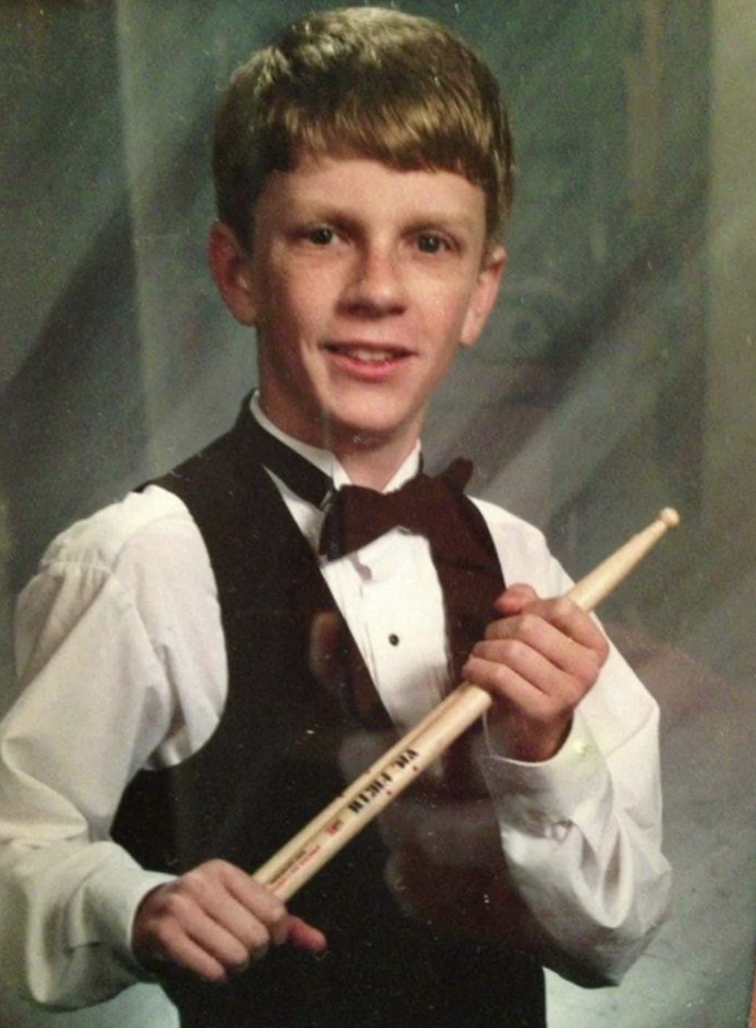 A young person with short hair is dressed in formal attire: a white dress shirt, a black bow tie, and a black vest. They hold a drumstick in one hand and pose with a smile against a neutral background.