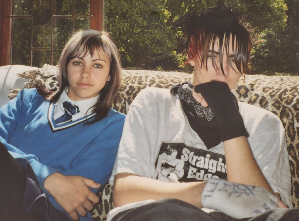 Two teenagers sit on a patterned couch. The girl on the left has dyed hair, wears a blue sweater over a collared shirt with a necktie. The boy on the right has spiked hair dyed red at the ends, wears a printed T-shirt, a bandana around his neck, and fingerless gloves.