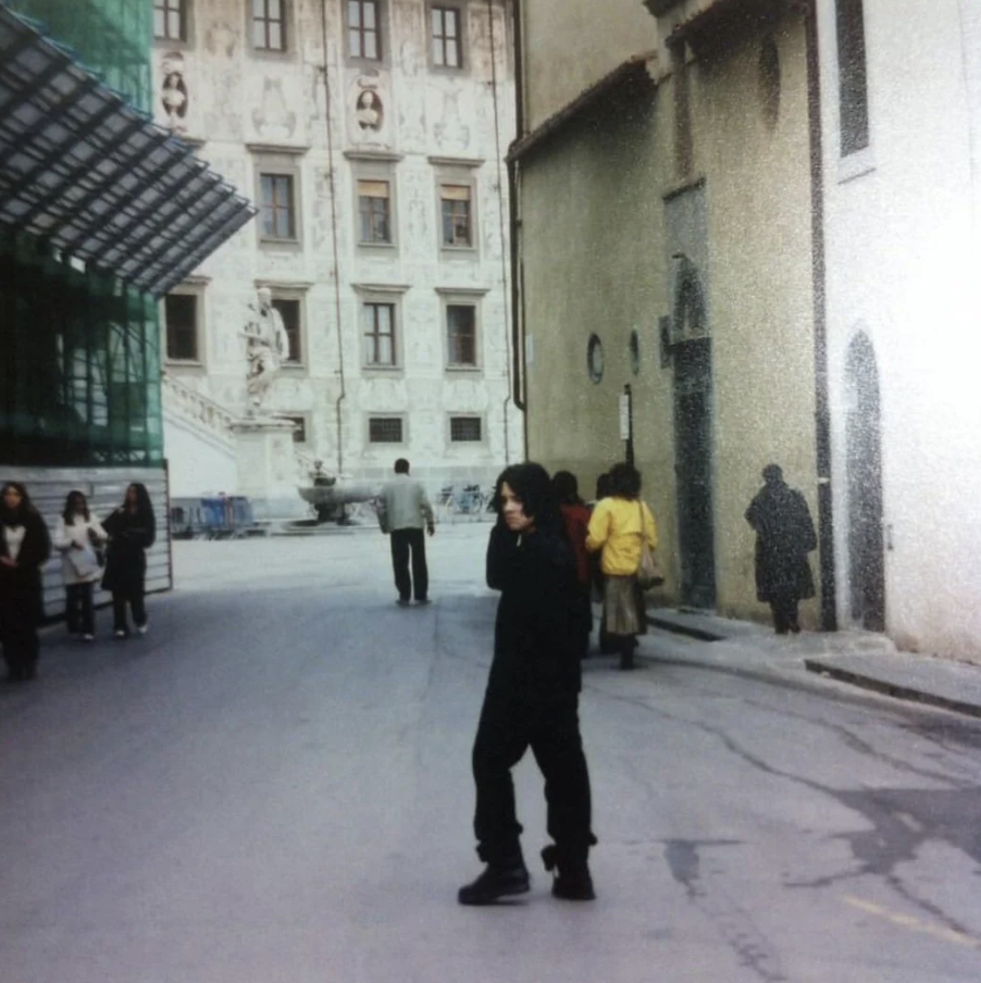 A person dressed in black walks in a European city square with historical buildings and statues in the background. A group of people can be seen walking and standing around. The atmosphere appears to be calm and overcast.