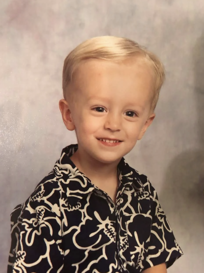 A young child with a big smile wearing a black shirt with a white floral design poses for a portrait photo in front of a neutral, light-colored background. The child has light blonde hair and is looking straight at the camera.