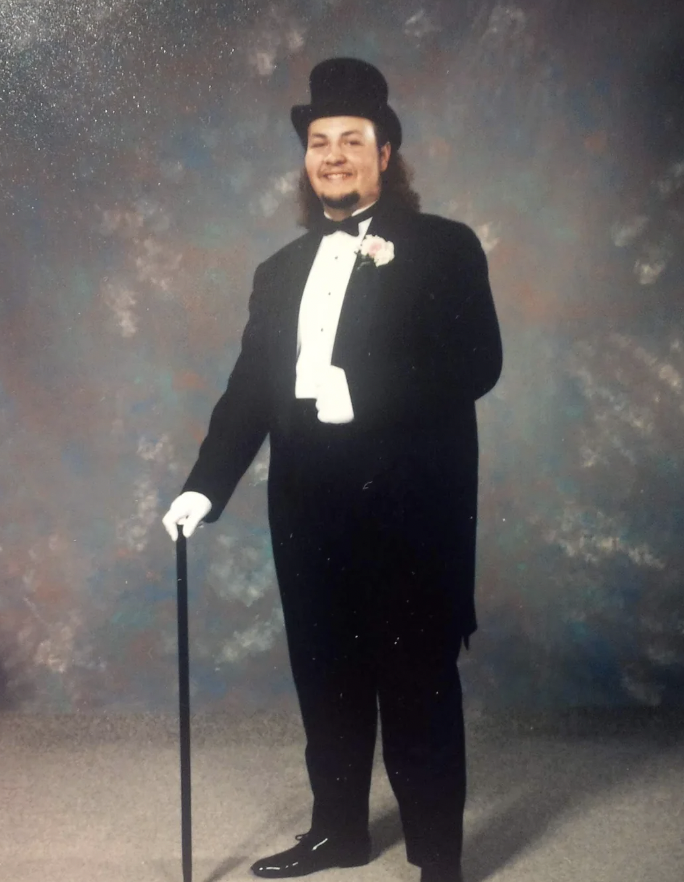 A man is dressed formally in a black tuxedo with a white shirt, black bow tie, and a top hat. He is smiling and holding a black cane in his gloved hand. The background is a simple, mottled pattern.