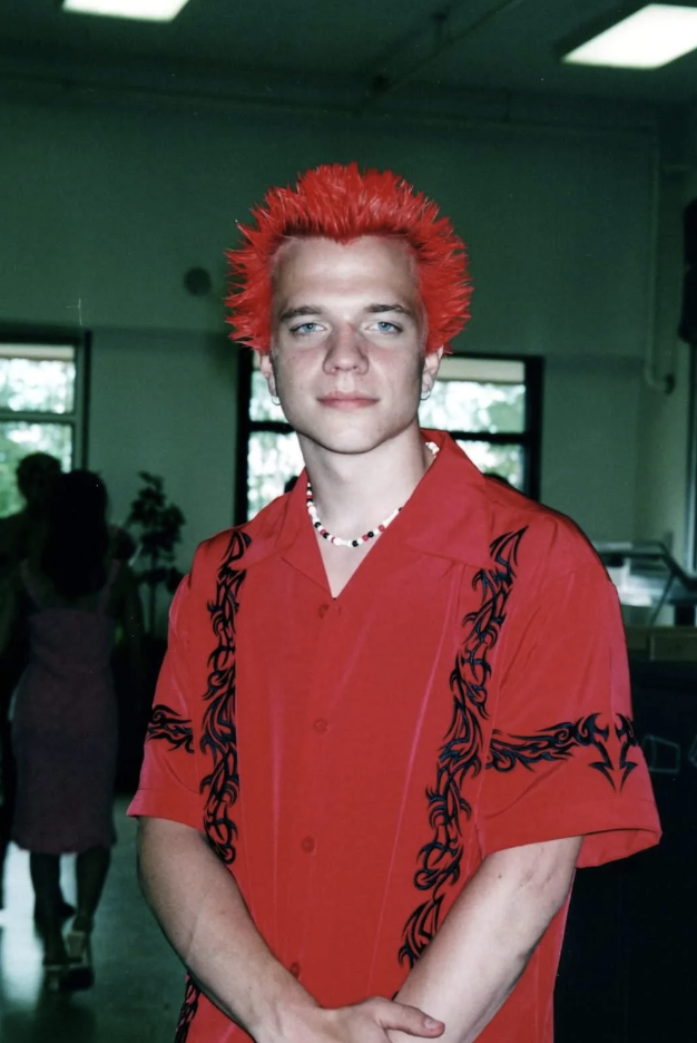 A person stands indoors with bright red spiked hair, wearing a red shirt adorned with black tribal patterns. They are looking directly at the camera with a neutral expression. The background includes people and windows letting in natural light.