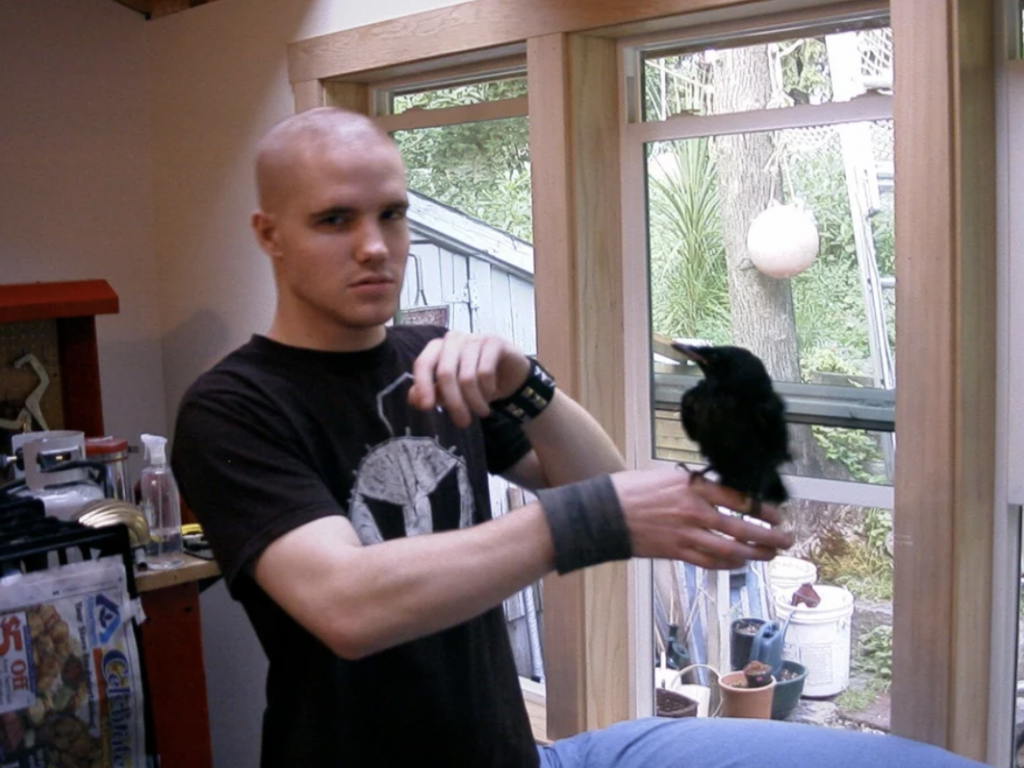 A man with a shaved head and wearing a black t-shirt stands in a room with large windows, holding a black bird on his right hand. The room has various items on shelves and a window showing an outdoor area with trees and plants.