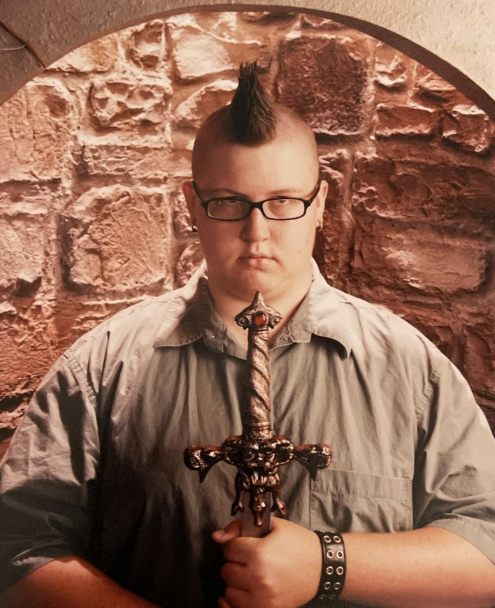 A person with a mohawk haircut and wearing glasses stands in front of a stone wall, holding a decorative sword upright. The individual is dressed in a button-up shirt and a studded bracelet. The expression is serious and focused.