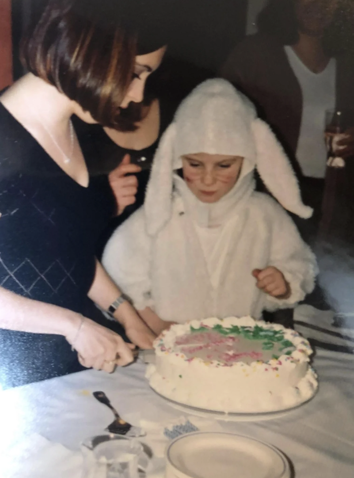 A child dressed in a white bunny costume stands at a table, looking at a cake while a woman beside them cuts a slice. The cake is decorated with green and pink icing and colorful sprinkles. There are other people in the background.