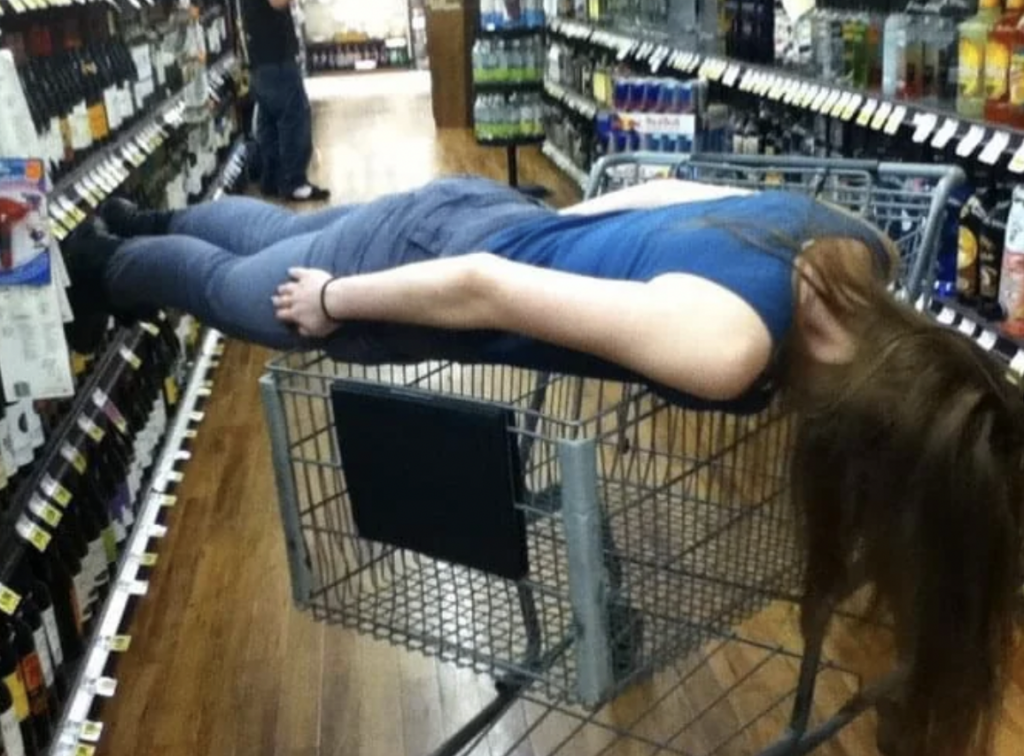 A person with long hair, wearing a blue shirt and gray pants, is lying face-down (planking) across a shopping cart in a grocery store aisle. The setting features shelves stocked with various bottles, and another person stands in the background.