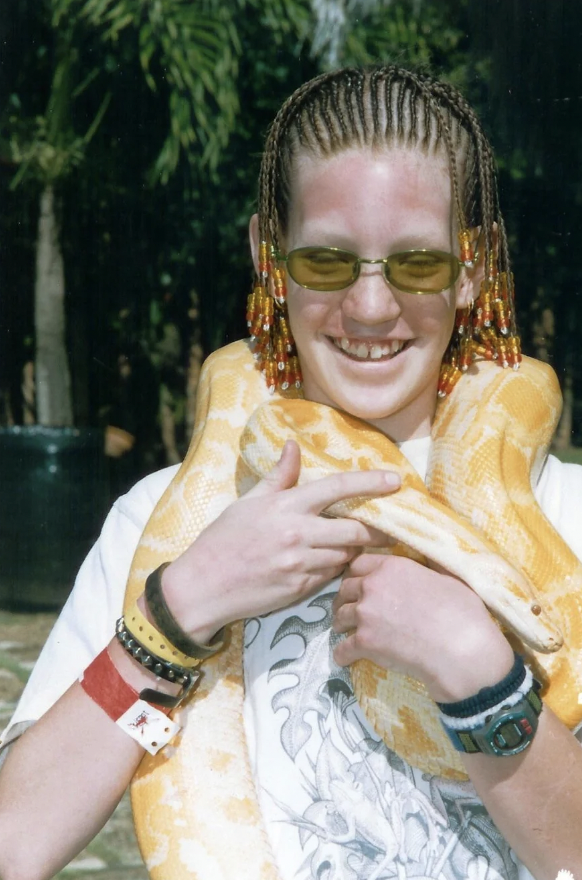 A person with braided hair and wearing sunglasses smiles while holding a large yellowish snake draped around their neck. The person is outdoors, wearing a white shirt and several bracelets on their left wrist. Trees and greenery are visible in the background.