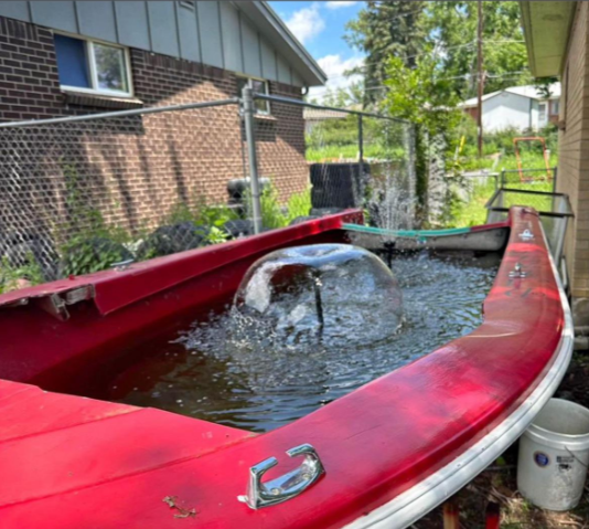 A red boat has been converted into a small backyard fountain, filled with water and a fountain jet spraying water upward. The boat is situated beside a brick building, with fencing and greenery in the background.