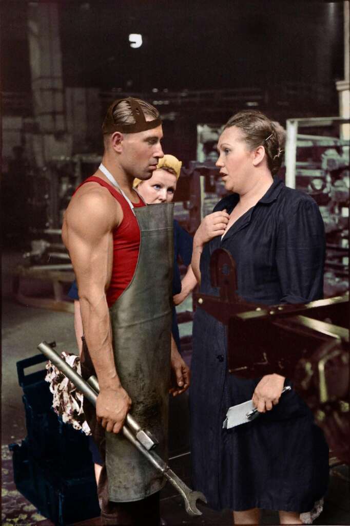 A sepia-toned image featuring a muscular man in a red tank top and grey apron holding a large wrench, looking towards a woman in a dark dress holding a clipboard. Another woman with a headscarf peeks from behind the man's shoulder in an industrial setting.