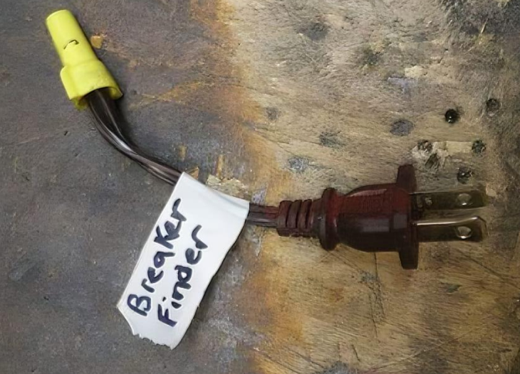 A homemade electrical device labeled "Breaker Finder" shows a three-pronged plug spliced into a single wire. The wire is capped with a yellow wire nut. The device is placed on a stained and worn wooden surface.