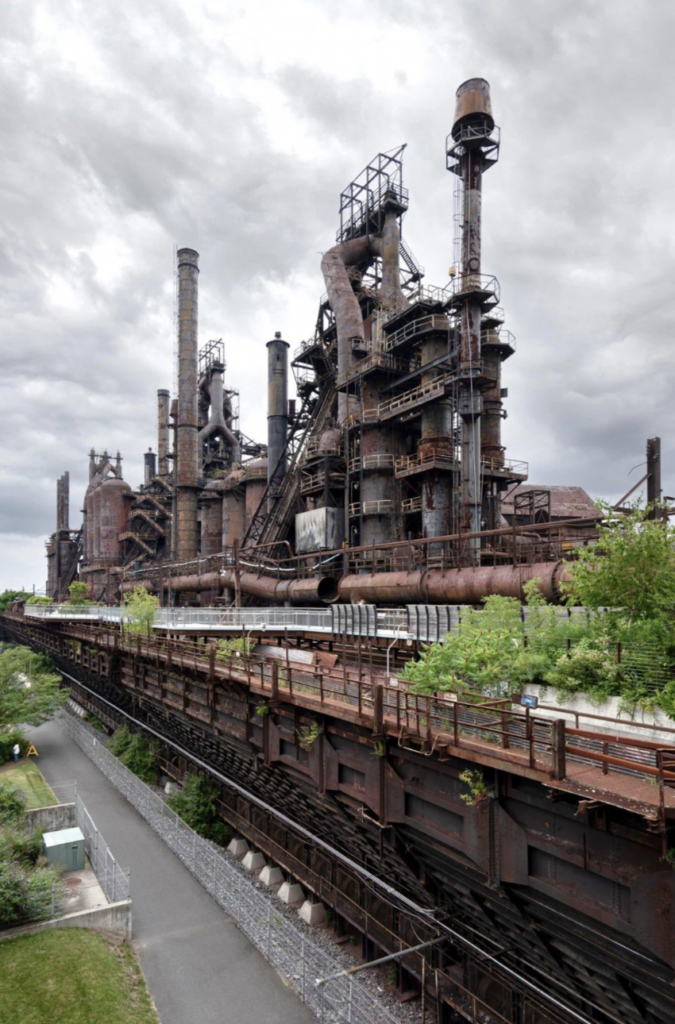 An old, abandoned industrial complex featuring rusted smokestacks and pipes. Overgrown vegetation is reclaiming parts of the structure, while a pathway runs parallel to it below. The sky is overcast, adding a gloomy atmosphere to the scene.