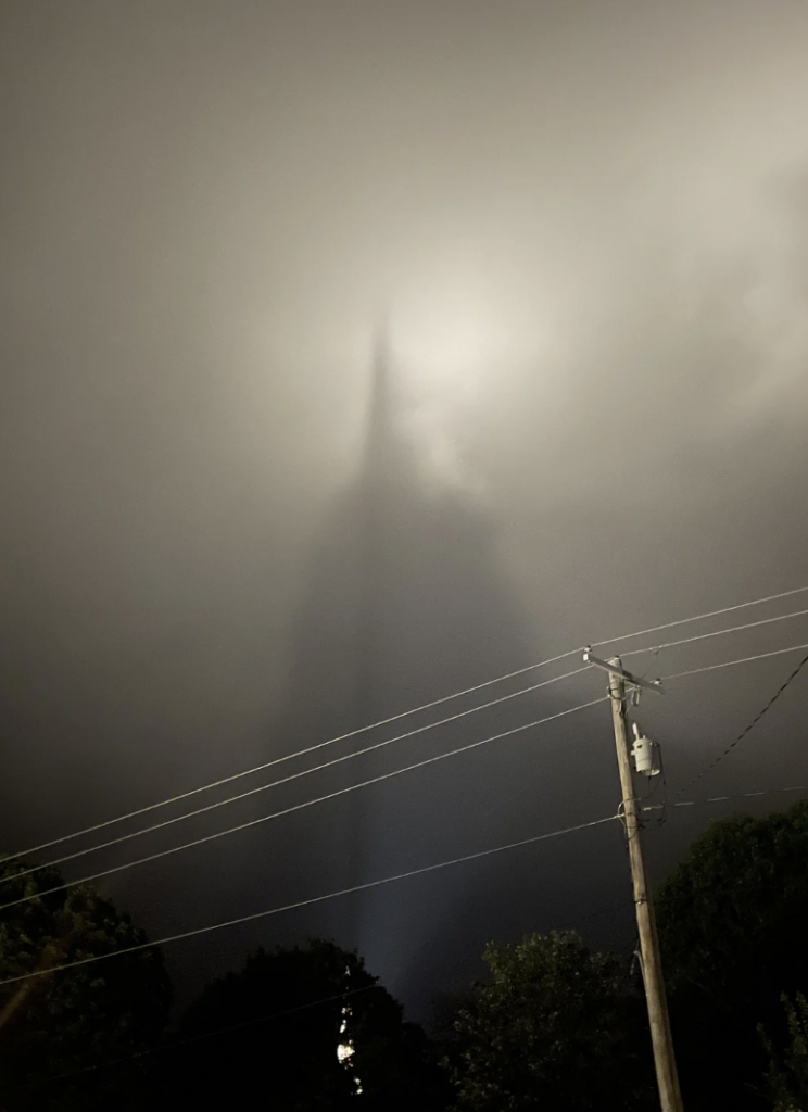 A tall structure, possibly a tower or skyscraper, is seen in a misty, foggy environment with a bright light at its peak. The image is dark and the structure casts a shadow through the fog. Trees and a utility pole with power lines are visible in the foreground.