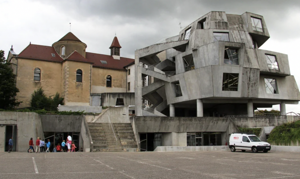 A group of people walks towards a unique, modern concrete building with angular shapes and irregular windows. Adjacent is an older stone building with a bell tower and red roof. A white van is parked on the right in a large, empty paved area.