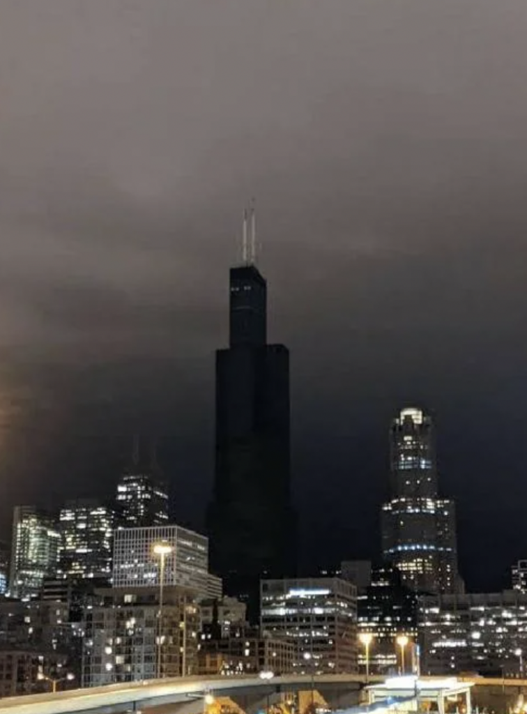 A nighttime cityscape featuring a skyline with tall buildings under a cloudy sky. The tallest building in the center dominates the view, with smaller illuminated buildings surrounding it. Streetlights and lighted windows create a contrast with the dark sky.