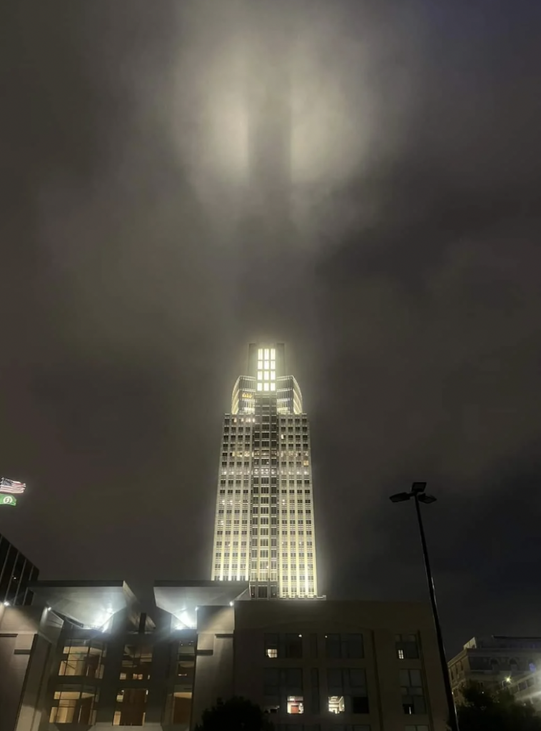 A tall building illuminated at night with lights shining upward, creating an ethereal glow against a cloudy sky. The building's design features illuminated windows and a spire-like top, making it stand out in the nighttime environment.