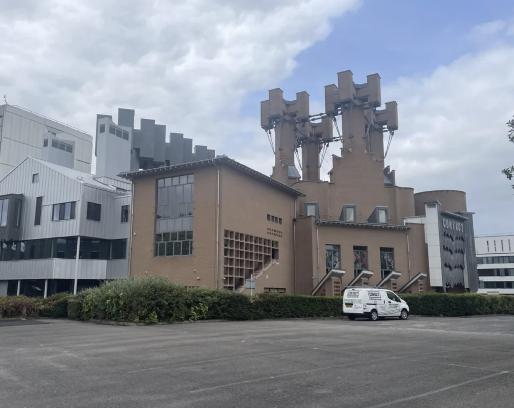 A modern industrial building with a unique brown brick structure featuring large external pipes and chimneys. Adjacent to it is a white and gray building with multiple windows. A white vehicle is parked in the empty lot in front. Cloudy sky overhead.