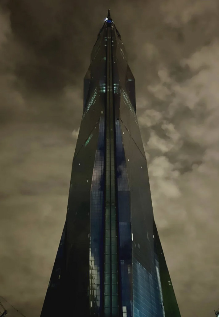 A tall, dark skyscraper extends into a cloudy night sky, illuminated by small, scattered lights on its surface. The building's reflective glass exterior captures the dramatic, cloud-filled atmosphere above.