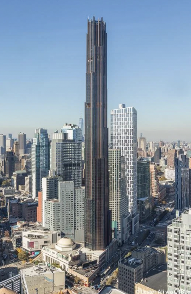 A tall, dark skyscraper towers over a densely built urban area in downtown Brooklyn. The building has a sleek, modern design and is surrounded by shorter, diverse-looking buildings under a clear blue sky.