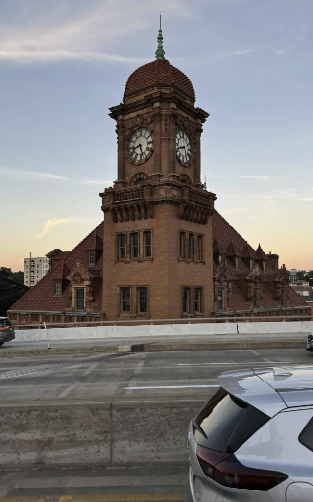 A historic, red-brick building with a prominent clock tower stands in front of a clear evening sky. The building features ornate detailing and a steep, triangular roof. Part of a road and the rear of a car are visible in the foreground.