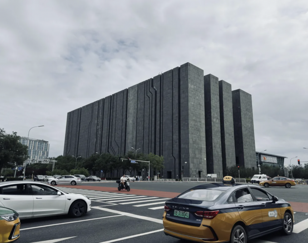 A modern, large, rectangular building with a dark exterior stands under a cloudy sky at a street intersection. Various vehicles, including cars and taxis, are seen waiting at the traffic lights in the foreground.