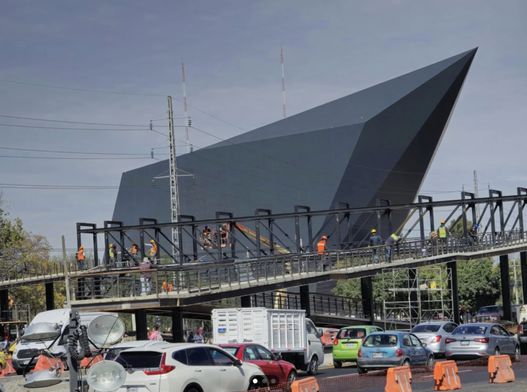 Construction workers are standing on a partially completed pedestrian bridge above a busy intersection filled with various cars. In the background, there's a modern angular building with a dark facade and sharp edges.