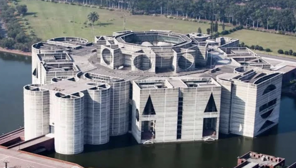 Aerial view of a large, modern architectural building with geometric shapes and patterns. The structure is surrounded by water and greenery, with multiple circular and triangular designs visible on its facade. Trees and grass fields are in the background.