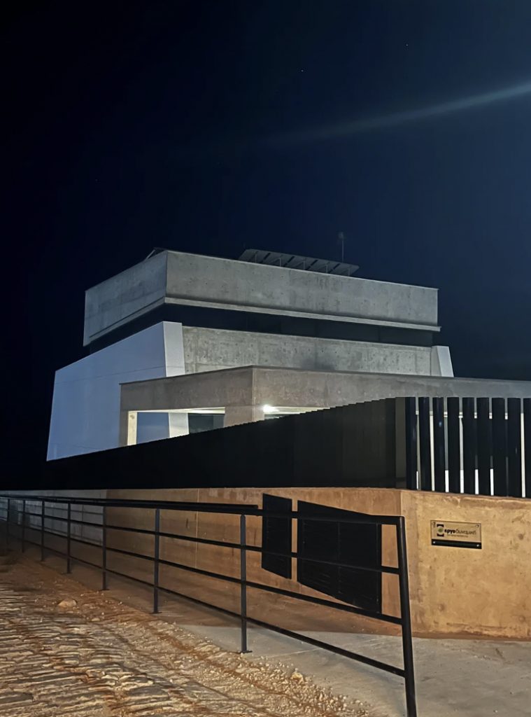 A modern, minimalist house with a flat roof sits illuminated under the night sky. The structure features a combination of concrete and white walls, with sleek lines and large windows. A black metal fence encloses the front yard, and a small sign is visible on the fence.