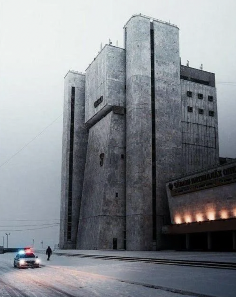 A modernist concrete building with tall, angular structures stands against a grey sky. A police car with flashing lights is parked on the snowy street in front. The scene is cold and desolate, with minimal activity and a subdued atmosphere.