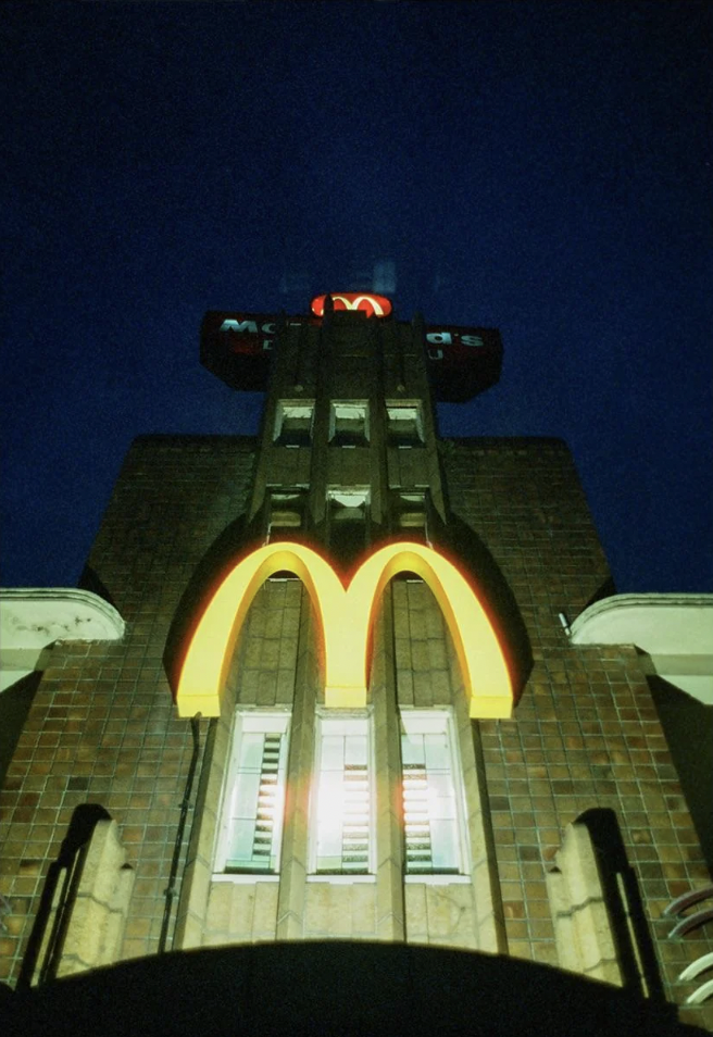 A McDonald's sign is illuminated at night on a tall, brick building. The iconic golden arches glow brightly, casting light on the structure with a deep blue night sky in the background.