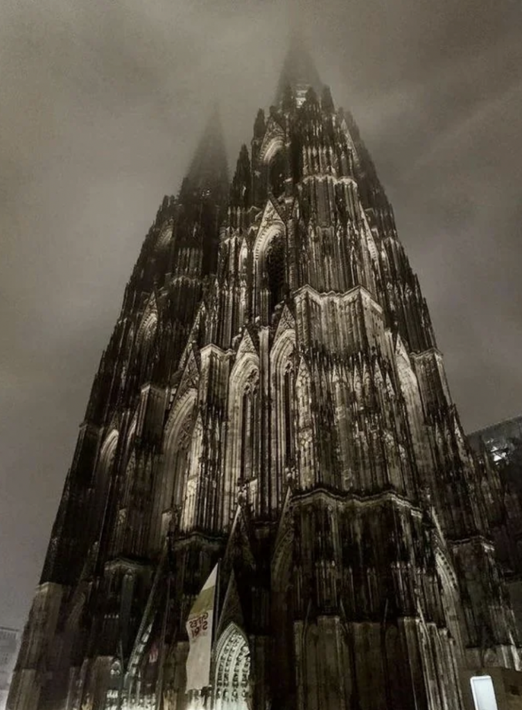 The image shows a gothic cathedral enveloped in fog at night. The structure is illuminated, highlighting intricate detailing and towering spires that seem to disappear into the misty sky above. The foreground includes faintly visible flags near the entrance.