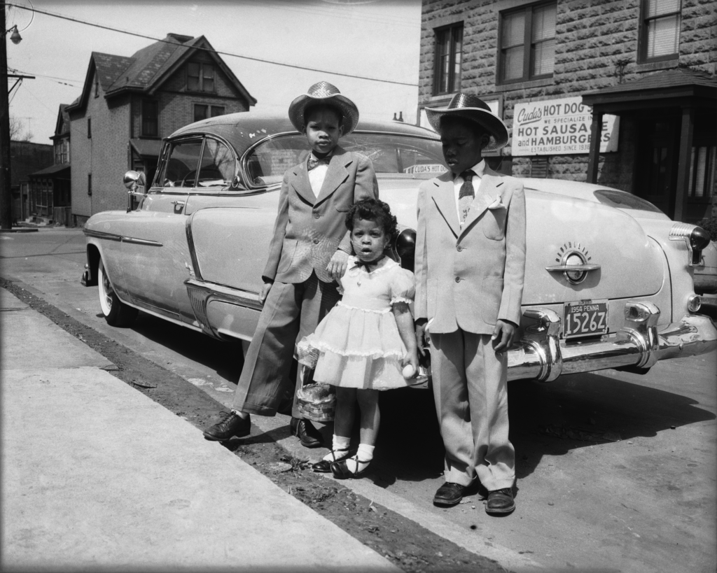 Three children stand beside a vintage car parked on a street. The two older boys wear suits and hats, flanking a younger girl in a dress and bow. A stone building with a sign reading "HOT DOGS and HAMBURGERS" is visible in the background.